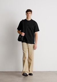 Μαύρο κοντομάνικο μπλουζάκι με κεντημένο λογότυπο, συνδυασμένο με μπεζ παντελόνι φαρδιάς γραμμής και μαύρα παπούτσια, κρατώντας τσάντα ώμου. Ουδέτερο φόντο.