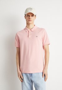 Junger Mann, der ein hellrosa Poloshirt, hellblaue Jeans und eine beige Kappe mit dem "GANT"-Logo trägt, steht vor einem einfarbigen weißen Hintergrund.