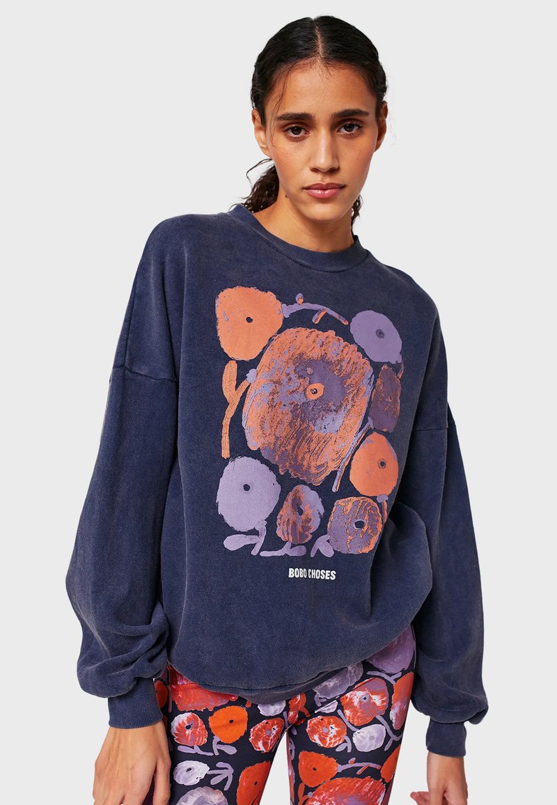 Nainen, jolla on yllään tummansininen huppari, jossa on suuria oransseja ja purppuranvärisiä kukkakuvioita, sekä yhteensopivat kukkakuosiset leggingsit.