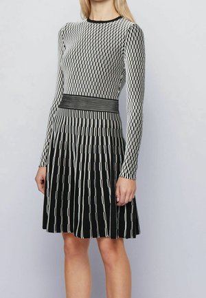 Mujer con un vestido de manga larga con estampado en blanco y negro, cintura ajustada y falda plisada, de pie contra un fondo claro y liso.