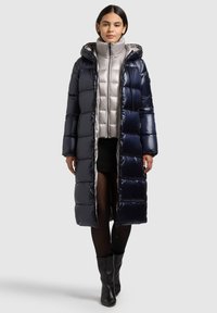 Hosszú, tengerészkék színű felfújt kabát fényes felülettel, kapucnival és kontrasztos ezüst belső béléssel. Texturált, steppelt mintázat.