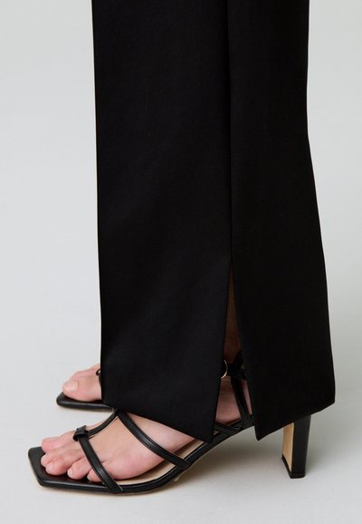 Ingrandimento di piedi che indossano sandali neri con tacco a fascia sotto pantaloni neri dritti con spacco laterale.