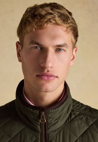 Junger Mann mit kurzen blonden Haaren und blauen Augen, der eine dunkelgrüne gesteppte Jacke trägt, die bis zum Hals zugezippt ist, schaut geradeaus.