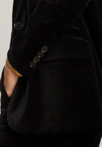 Blazer di velluto nero con cinque bottoni sul polsino. Presenta una tasca laterale e una fodera dorata visibile sulla manica sotto il polsino.