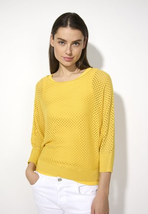 Gelber gestrickter Pullover mit lockerer Passform, ausgestattet mit einem Netzdesign und gerippten Bündchen, kombiniert mit weißen Hosen.