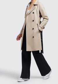 Beige trenchcoat med dubbelknäppt front, svarta knappar och sidofickor. Har ett bälte och kontrasterande svarta accenter; slät textur.