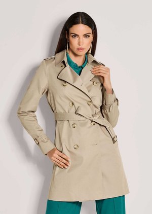 Frau trägt beige zweireihigen Trenchcoat mit Gürtel, türkisfarbenes Hemd und Hose, posiert mit der Hand nahe am Kragen vor einfachem Hintergrund.