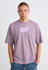 Mand med kort hår iført en oversized lyslilla T-shirt med tegneserie-tekst "RIPNDIP PUMA" og små hvide katteillustrationer, hænderne i lommerne.