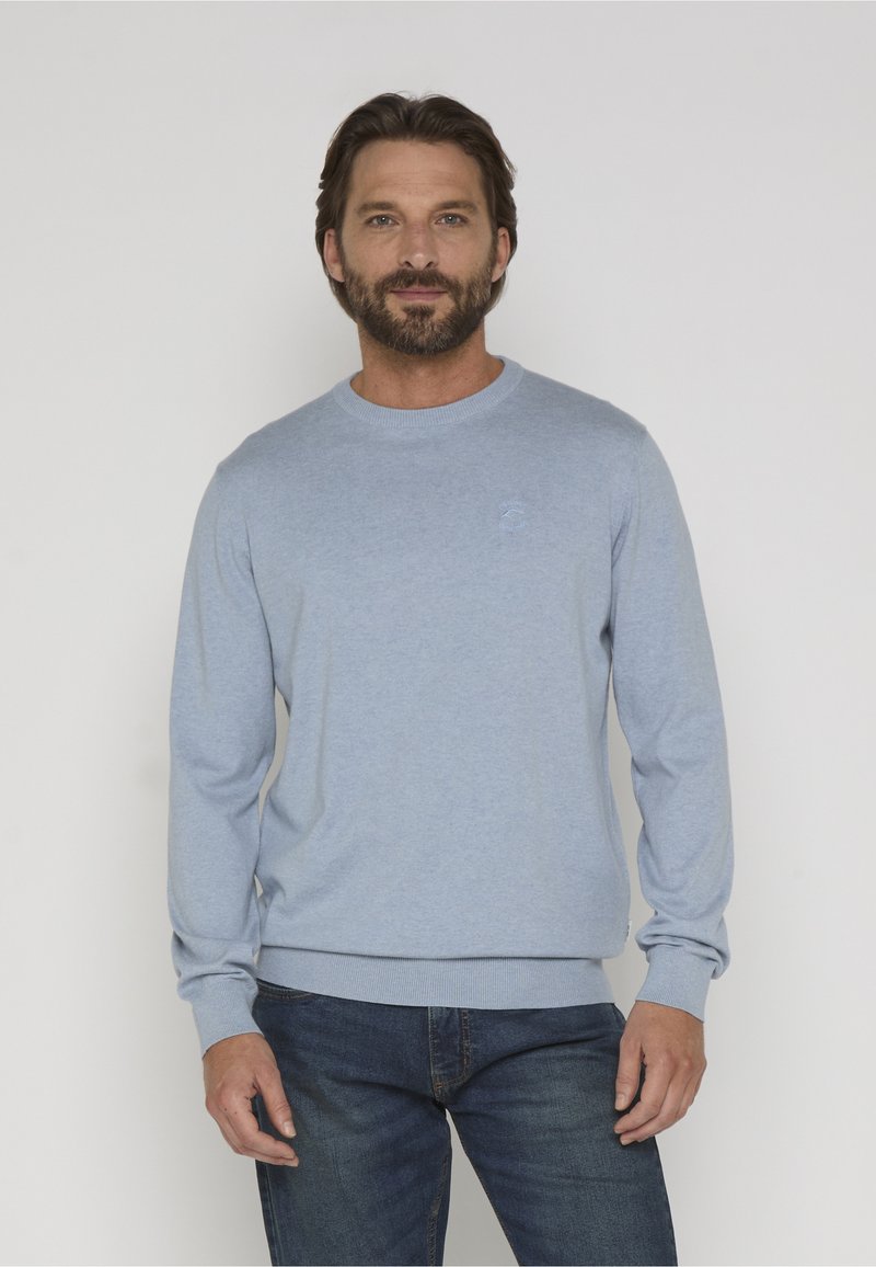 Homme barbu portant un pull ras du cou bleu clair et un jean bleu foncé, debout devant un fond clair uni.
