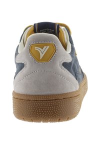 Zapatilla con una parte superior de ante gris y tela en azul marino, detalles en amarillo, suela de goma texturizada y un logo hexagonal en el talón.