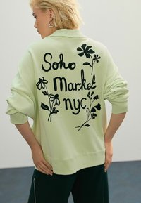 Hele roheline üleliigne spordipusa mustade lillemotiivide ja tekstiga "Soho Market nyc" seljal. Ribiga mansetid ja alumine äär. Üksik sobivus.