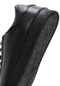 Black leather shoe with textured gray rubber sole and smooth upper. Features a rounded toe and decorative stitching along the edge.