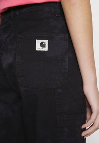 Personne portant un pantalon Carhartt noir avec une poche arrière visible et un patch de logo, à côté d'une partie d'une chemise rose et d'une main au repos.