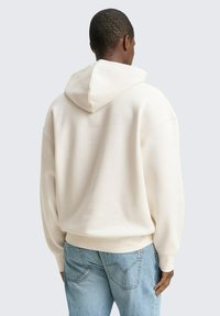 Haut en crème avec une grande capuche, des poignets et un ourlet côtelés ; porté avec un jean en denim bleu clair. Texture douce, coupe décontractée, design minimal.