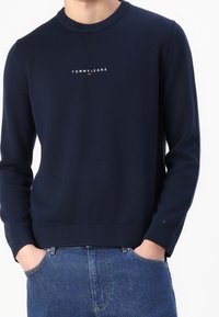 Granatowy sweter z dzianiny z prążkowanym dekoltem i dolnym wykończeniem, z białym logo "TOMMY JEANS" umieszczonym na środku klatki piersiowej. Standardowy krój, długie rękawy.