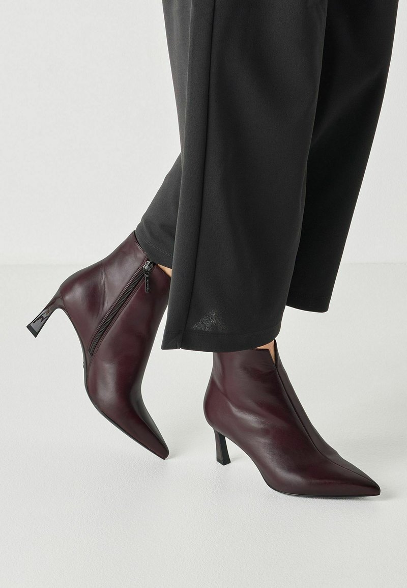 Personne portant des bottines bordeaux foncé à bout pointu avec des fermetures éclairs latérales et des talons de hauteur moyenne, associées à un pantalon large noir.