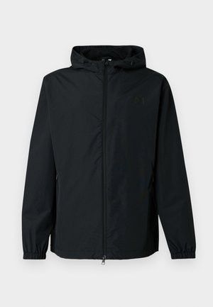 Črna vetrovka z zadrgo spredaj, nastavljivo kapuco, elastičnimi manšetami in diskretno podrobnostjo logotipa na prsih. Tkanina z teksturo.