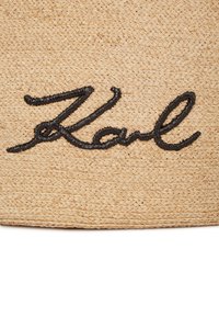 Vītā maisiņš no salmiem, brūnā krāsā, ar melnu izšūtu tekstu "Karl," izceļot gludu tekstūru un tīru dizainu.
