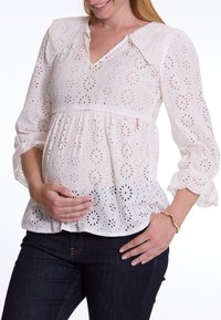 Witte blouse van oogjes met een V-hals, gerimpelde mouwen en een gathered taille. De stof heeft bloemen uitsparingen en is gecombineerd met donkere jeans.