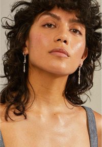 Pendientes de plata con piedras preciosas transparentes colgantes, llevados por un modelo con cabello rizado, que muestra una textura de piel suave y un maquillaje natural.