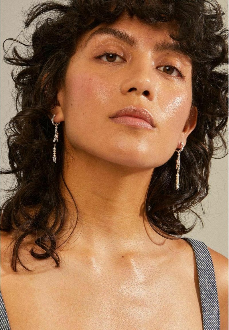 Silver earrings with dangling clear gemstones, worn on a model with curly hair, showcasing a smooth skin texture and natural makeup.