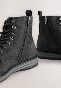 Schwarze Wildleder-Stiefeletten mit strukturierter Oberfläche, Schnürung vorne, Seitlichem Reißverschluss und Gummisohle mit Profilmuster.