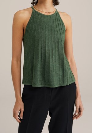 Vrouw draagt groene mouwloze ribgebreide haltertop en zwarte broek, met gouden ketting, staand tegen een effen achtergrond.