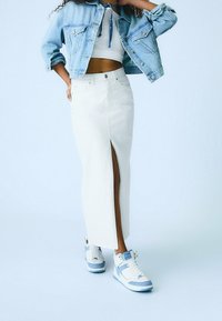Donna con giacca di jeans azzurro chiaro, top corto bianco, lunga gonna bianca con spacco frontale e scarpe da ginnastica bianche con dettagli azzurri.
