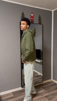 Veste vert olive avec capuche et poche avant, portée sur un jean bleu clair déchiré, debout à côté d'un miroir en pied.