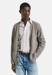 Junger Mann in einer grauen Knopfjacke über einem weißen Hemd mit roten Streifen und hellblauen Jeans, mit der Hand in der Tasche, neutraler Hintergrund.