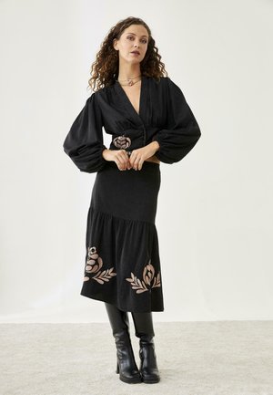 Mujer con cabello rizado que lleva un vestido negro con mangas abullonadas, bordado floral en la cintura y el dobladillo, de pie con botas negras hasta la rodilla.