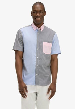 Homme souriant portant une chemise boutonnée à manches courtes avec des panneaux à carreaux bleus et gris, col et poche roses, debout les mains détendues.