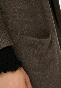 Brązowy dziany kardigan z przednią kieszenią i teksturowanym, czarnym, warstwowym mankietem. Materiał jest miękki z delikatnym połyskiem.
