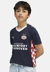 Tamno plavi nogometni dres s bijelim ovratnikom, logotipom i crvenim kariranim uzorkom sa strane. Ima kratke rukave i prozračnu tkaninu.