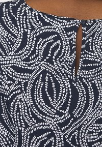 Mörkblå blus med vitt abstrakt mönster, med ett nyckelhål i ryggen och en ensam knapplåsning. Slät, lättviktig tyg.