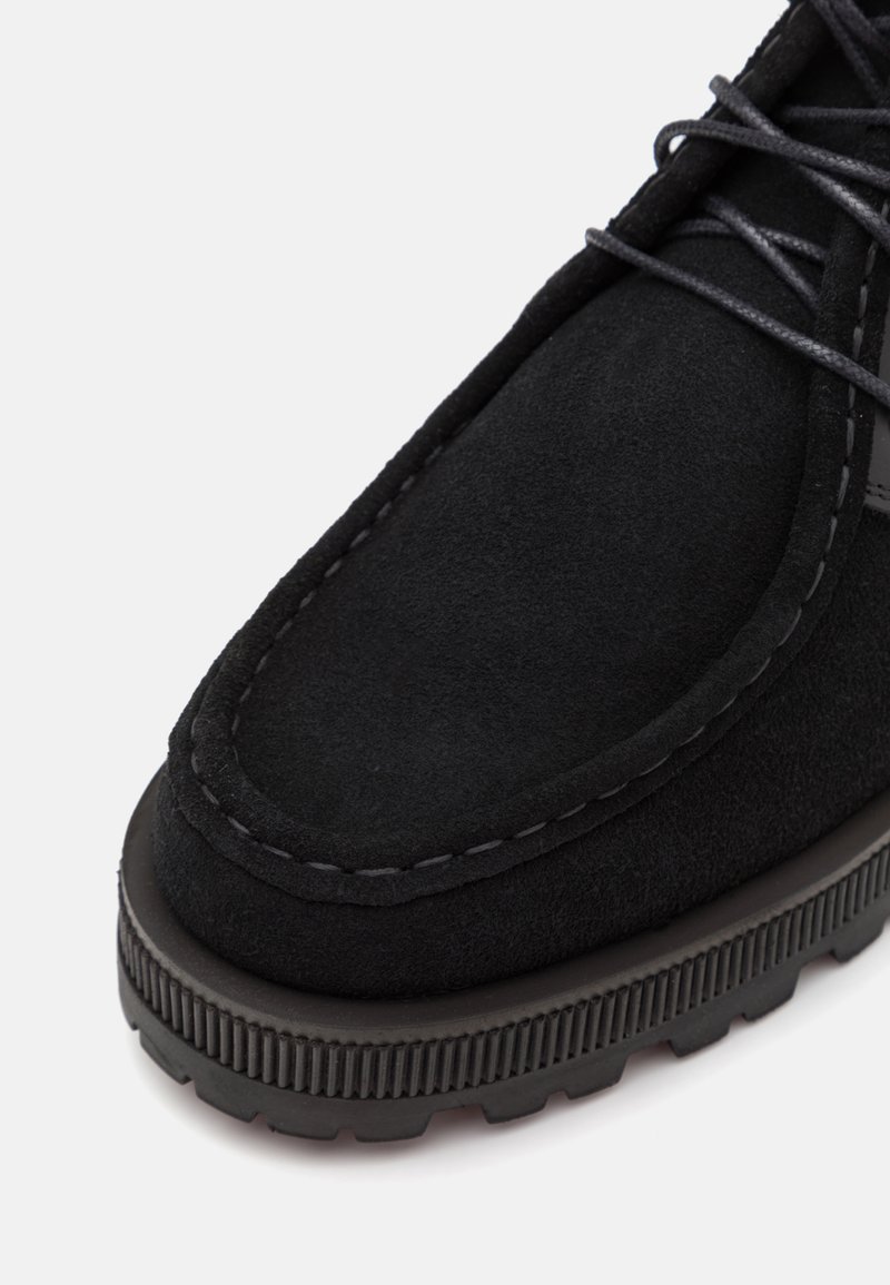 Chaussure en suede noir avec un bout rond, des coutures contrastées et une semelle en caoutchouc texturé avec des rainures pour une meilleure adhérence.