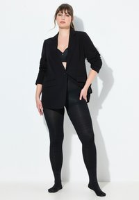 Blazer nero su misura con chiusura frontale a bottoni e due tasche, abbinato a collant neri opachi con un leggero motivo.
