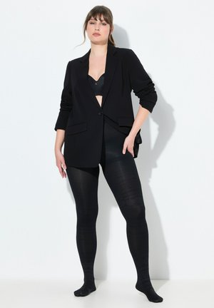 Zwarte op maat gemaakte blazer met een sluiting met knopen aan de voorkant en twee zakken, gecombineerd met zwarte opaque panty’s met een subtiel patroon.