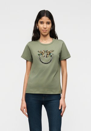 Kvinde iført olivengrøn t-shirt med palietfugl-design og mørkeblå jeans, stående mod en ensartet lysegrå baggrund.