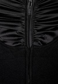 Lähikuva mustasta rypistetyistä kankaasta, joka on ribatusta neuloksesta, keskellä vetoketju, jossa on metallinen vetotappi, johon on kaiverrettu "MORGAN."