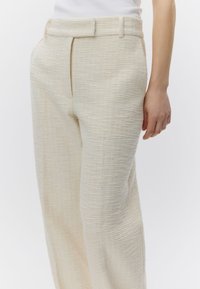 Gros plan sur une personne portant un pantalon taille haute texturé de couleur crème avec des poches latérales et un haut blanc rentré.