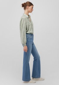Green patterned blouse with a collar, long sleeves, and gathered cuffs, paired with light blue flared jeans and white sneakers.