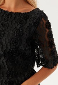 Sort blomster-lace top med korte ærmer, der har lagdelte stofroser og en gennemsigtig tekstur. Høj halsudskæring, elegant design, intrikate detaljer.