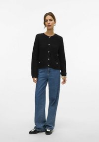 Cardigan nero a maglia con tessuto testurizzato e bottoni dorati, abbinato a jeans blu a gamba larga. Il modello mostra un taglio rilassato.