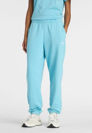 Persoon draagt lichtblauwe joggingbroek met elastische boorden en witte sneakers, staand tegen een effen witte achtergrond.