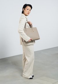 Sac fourre-tout texturé beige avec doubles poignées supérieures, quincaillerie dorée et forme structurée, présenté par un mannequin dans un costume de couleur claire.