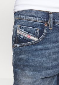 Tmavě modré džínové kalhoty s opranou texturou, s nášivkou s logem "Diesel Industry" na zadní kapse a měděnými kovovými doplňky.