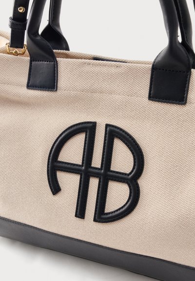 Μπεζ τσάντα χειρός από καμβά με μαύρες δερμάτινες λαβές, περιμετρικό μαύρο τελείωμα στον πάτο και μαύρο κεντητό λογότυπο "AB" στο μπροστινό μέρος.