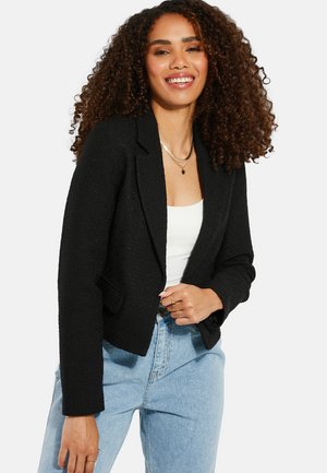 Schwarze, strukturierte Blazerjacke mit tailliertem Schnitt, Ein-Knopf-Verschluss und zwei Fronttaschen. Kombiniert mit einem weißen Oberteil und hoch taillierten blauen Jeans.