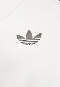 Juodas siuvinėtas Adidas trefoil logotipas, centruotas ant balto audinio fono.